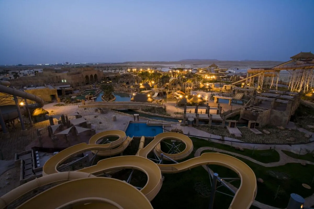 At dusk looking down from atop a yellow winding waterpark slide, over a well-lit path including other slides and pools