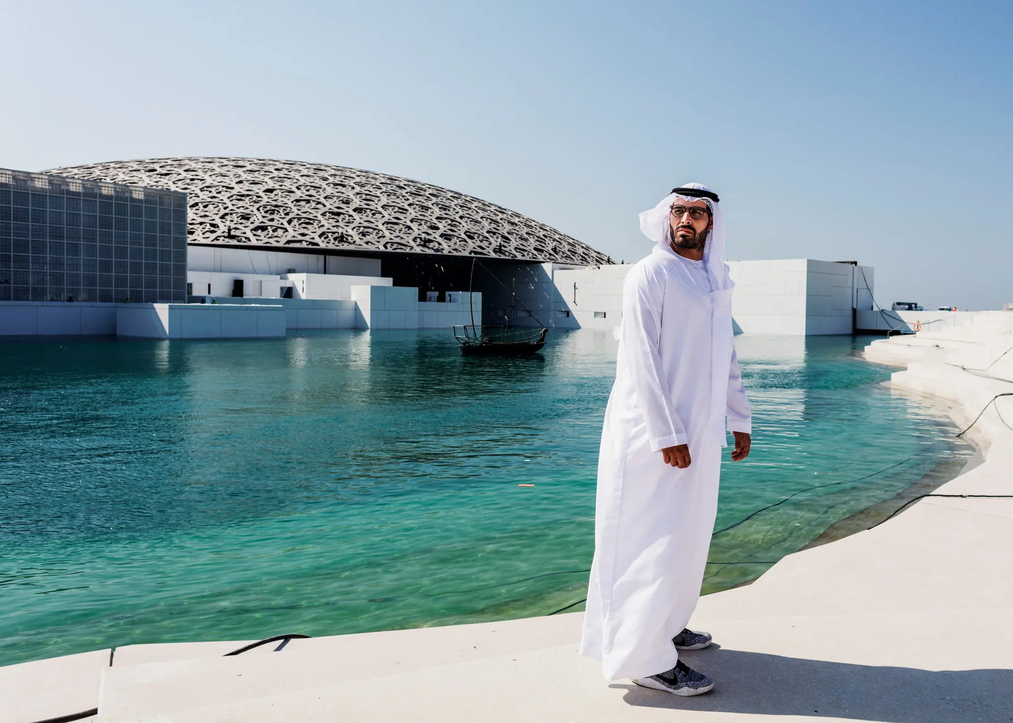A man dressed in traditional white Middle Eastern attire stands near a body of water with the modern dome-shaped structure of the Louvre Abu Dhabi visible in the background. The building's intricate lattice design contrasts with the clear blue sky and the turquoise water surrounding it.