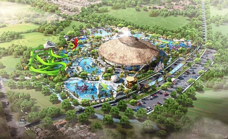 A waterpark is surrounded by lush green fields and trees. To the left side of the park is a twisting and turning bright green water slide. The center of the park features a Hawaiian style building, with multiple pools surrounding the building