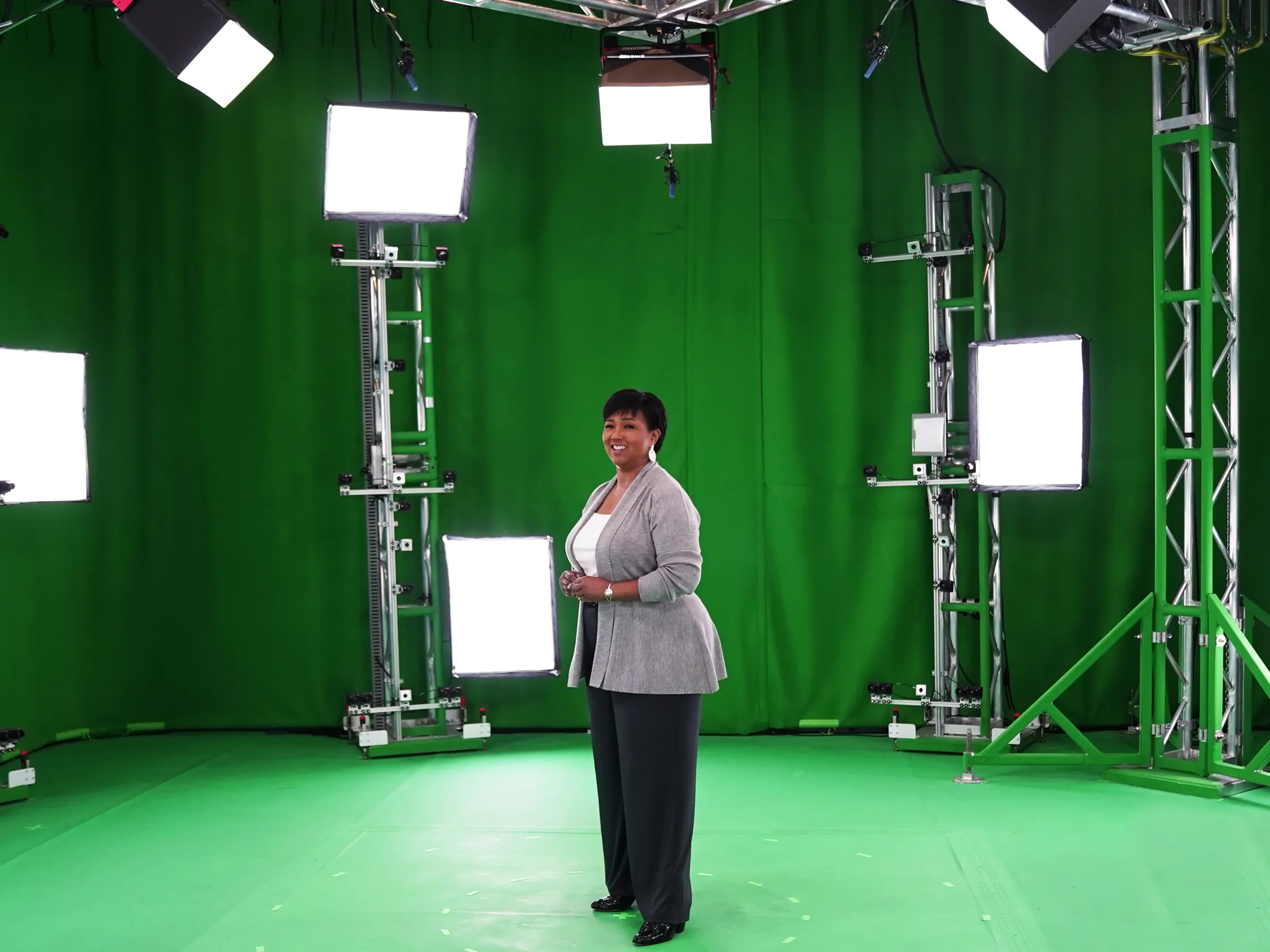 Green Screen room, arranged with several light boxes and cameras, strewn around a smiling woman in business casual clothing